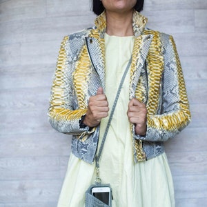 May include: A snakeskin-patterned jacket with yellow and gray tones, featuring a zipper closure and a collared neckline. A gray phone case with a strap is hanging from the neck. The person is wearing a light yellow dress.
