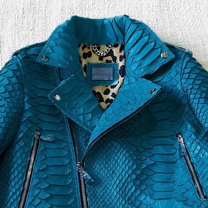 以下が含まれることがあります： ヘビ柄のターコイズブルーの革ジャン。ジッパー留めと襟付きです。