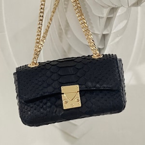 Peut inclure: Un sac à main en cuir de serpent noir avec une bandoulière en chaîne dorée et un fermoir doré. Le sac est suspendu à une sculpture de tête de cerf blanche.