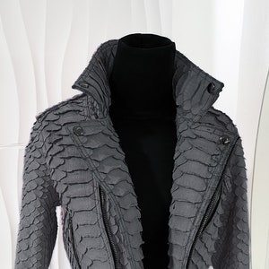 以下が含まれることがあります： ユニークな鱗模様のグレーのテクスチャード加工ジャケット。 クロップドジャケットは、ハイカラー、非対称ジッパー開閉、袖にジッパーが特徴です。 ジャケットは黒いマネキンに展示されており、そのデザインと詳細が示されています。