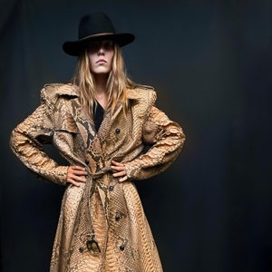 May include: A woman wearing a long, brown snakeskin trench coat with a black hat and black heels.
