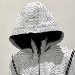 以下が含まれることがあります： 蛇柄の白い革のジャケットで、フード付き。ジッパーで閉めることができ、ポケットが2つある。