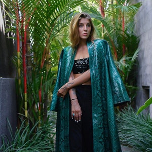 May include: A woman wearing a long, green snakeskin patterned jacket with a black top and black pants. She is standing in a tropical garden setting.