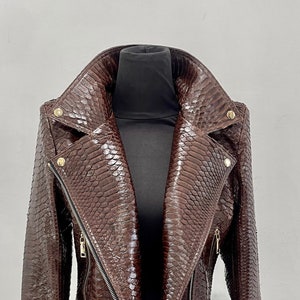 May include: A brown leather jacket with a snakeskin pattern. The jacket has a zipper closure, a belt, and gold-toned hardware.