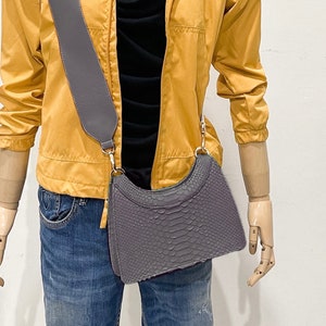 May include: A grey snakeskin crossbody bag with a grey adjustable strap and a gold buckle. The bag is worn over a yellow jacket and blue jeans.