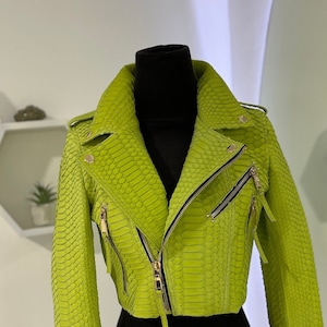 以下が含まれることがあります： 明るい緑色の革のジャケットで、ジッパー留めと金色の金具が付いています。ジャケットはヘビ革の模様で、ウエストが短くなっています。