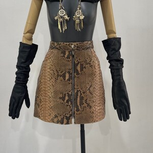 May include: A black leather beret with a snakeskin pattern, a black leather mini skirt with a zip down the front, and black leather gloves. The mannequin has blue hair and is wearing a silver chain necklace with charms.