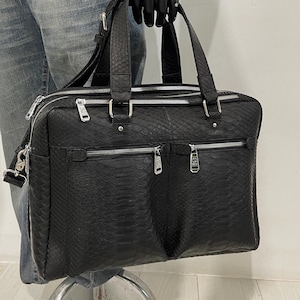 以下が含まれることがあります： 蛇柄の黒い革製のブリーフケース。シルバーの金具が付いています。トップハンドル、ショルダーストラップ、複数のジッパー付きコンパートメントがあります。