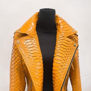 以下が含まれることがあります： ジッパー留めとベルトが付いた黄色のヘビ革ジャケット。ジャケットはスタンドカラーと長袖です。