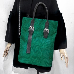 以下が含まれることがあります： 黒い革のストラップとバックルが付いた緑色のヘビ革のトートバッグ。バッグにはシルバーのチェーンストラップが付いており、黒いコートと黒いベースボールキャップを着たマネキンが持っています。