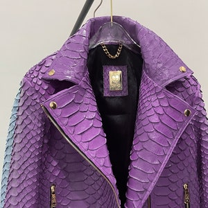 以下が含まれることがあります： 紫色のヘビ革のジャケットで、ジッパー留めと金色の金具が付いています。ジャケットはスタンドカラーで、前面にジッパー付きのポケットが2つ付いています。