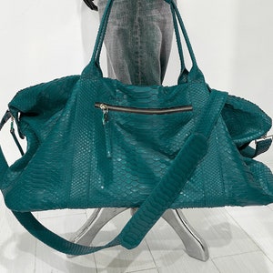 May include: A teal green leather duffel bag with a long shoulder strap. The bag has a zipped pocket on the front and is made of a textured material.