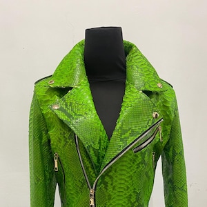 May include: A vibrant green snakeskin-textured jacket with a classic biker style. Features a notched collar, asymmetrical zipper closure, and gold-tone hardware. The jacket is displayed on a black mannequin, showcasing its cropped length and fitted silhouette.