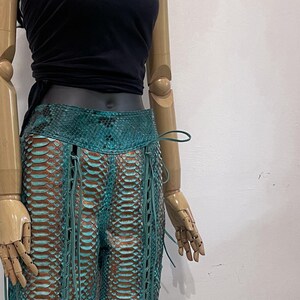 以下が含まれることがあります： ターコイズ色のヘビ革のパンツで、レースアップのデザインになっています。パンツはフレアになっていて、ハイウエストです。
