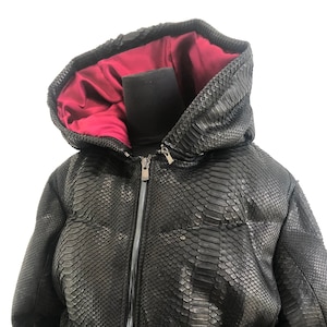 May include: A black snakeskin puffer jacket with a red lining and a zip closure. The jacket has a hood and a drawstring at the waist.