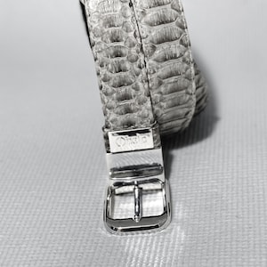 May include: A silver belt buckle with the word "Orisha" engraved on it. The belt is made of light gray snakeskin.