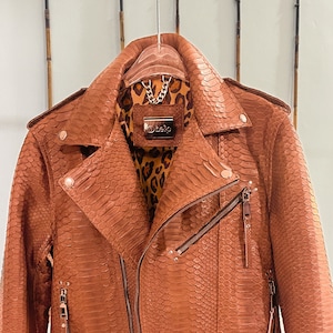 May include: A brown leather jacket with a snakeskin pattern. The jacket features a zip fastening, a belt at the waist, and a leopard print lining. The label on the jacket reads "Belpo".