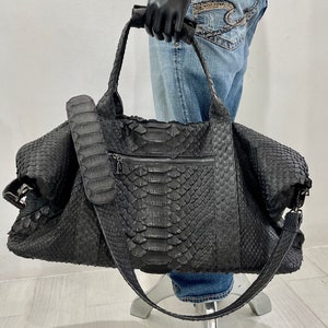 May include: A black leather travel bag with a snakeskin pattern. The bag has a top handle, a shoulder strap, and a zipped pocket on the front.