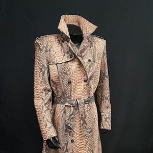 以下が含まれることがあります： ダブルブレストの留め具、ベルト、ハイカラーが付いた、長く、茶色と黒のヘビ革のトレンチコートです。