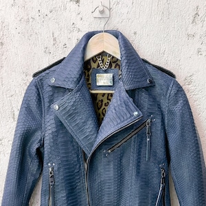 以下が含まれることがあります： ジッパー留めとウエストにベルトが付いた、青いヘビ革のバイクジャケットです。