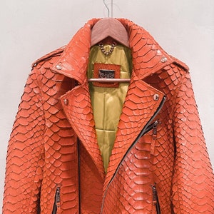 以下が含まれることがあります： 明るいオレンジ色のヘビ革のジャケットで、ジッパー留めとベルトが付いています。ジャケットは緑色の裏地と「DEBI」と書かれたラベルが付いています。