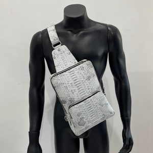 以下が含まれることがあります： 白いヘビ革のショルダーバッグ。ジッパー付きポケットとシルバーのジッパーが付いています。