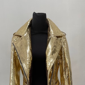 以下が含まれることがあります： ヘビ柄の金色の革ジャンパー。ジッパー留め、ベルト、襟付きです。