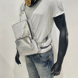 May include: A silver snakeskin-embossed crossbody bag with a black zipper and strap. The bag is worn over a white t-shirt and blue jeans.