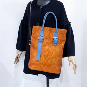 以下が含まれることがあります： 青いストラップとバックルが付いた茶色の革製のトートバッグ。バッグはヘビ柄で、黒いコートと黒い野球帽をかぶったマネキンが持っています。