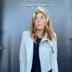 May include: A white leather jacket with a snakeskin pattern. The jacket has a zipper closure and a collar that can be worn up or down. The model is wearing a black top and a green and white patterned headband.