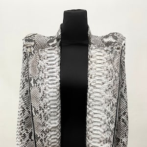 以下が含まれることがあります： グレーとホワイトのヘビ柄のジャケットで、ジッパーで閉まります。ジャケットはスタンドカラーで、少し丈が短くなっています。