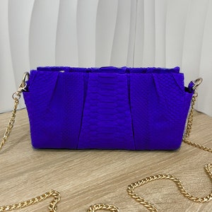 May include: A small, bright purple clutch bag with a gold chain strap. The bag has a textured, snakeskin-like pattern.