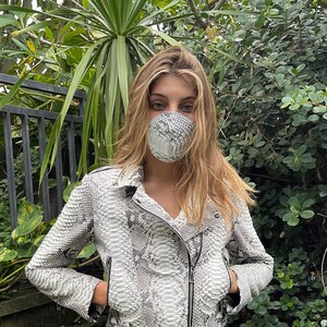 May include: A white and gray snakeskin jacket with a zipper closure and a belt. The jacket is cropped and has a relaxed fit. The model is wearing a white snakeskin face mask.