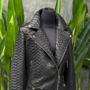 以下が含まれることがあります： ヘビ柄の黒い革のジャケットで、金色のバックルベルトが付いている。