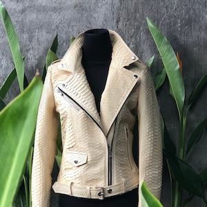 May include: A cream-coloured leather jacket with a snakeskin pattern. The jacket has a zip closure, a belt, and two pockets.