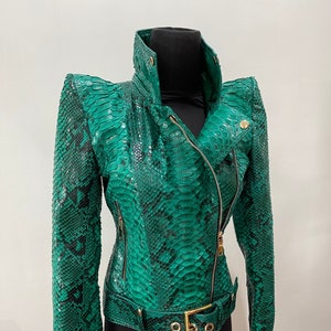 以下が含まれることがあります： 金色のジッパーとベルトが付いた緑色のヘビ革ジャケット。ジャケットはハイネックで、体にフィットしたシルエットです。