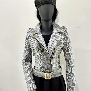 以下が含まれることがあります： 金色のジッパーとベルトが付いたグレーのヘビ革ジャケット。ジャケットはスタンドカラーと長袖です。