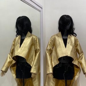 以下が含まれることがあります： ワニの型押し模様のゴールドメタリックジャケット。ジャケットはハイネックで、長く流れるようなバックスタイル。マネキンにディスプレイされています。