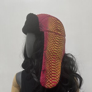 May include: A red and gold snakeskin trapper hat with a black faux fur brim. The hat has a long, pointed flap that hangs down the back of the neck.