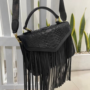 May include: Black leather handbag with a fringe detail and a snakeskin pattern. The bag has a top handle and a long shoulder strap.