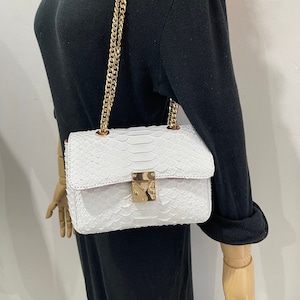 May include: White leather handbag with a snakeskin pattern and a gold chain strap. The bag has a gold lock and a pink lining.