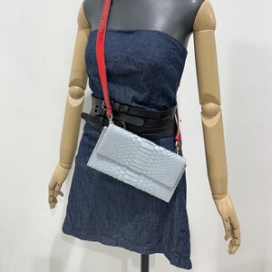 May include: A light blue snakeskin clutch bag with a red and black strap. The bag is worn over the shoulder of a mannequin.