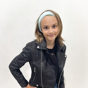 May include: A young person wearing a black leather jacket with a snakeskin pattern. The jacket has a zip fastening and a collar. The person is also wearing a light blue headband and blue denim shorts.