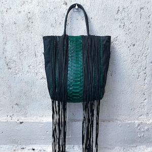 以下が含まれることがあります： 長いフリンジと緑色のヘビ革のパネルが付いた黒いトートバッグ。バッグはトップハンドルが付いており、フックに掛けられています。