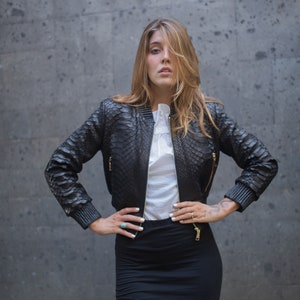 May include: A woman wearing a black leather jacket with a snakeskin pattern and a black mini skirt. She is standing in front of a grey wall.
