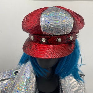 May include: A red and silver cap with a snakeskin pattern. The cap has a red brim and a silver, iridescent top section. Silver studs decorate the band. The cap is part of a fashion ensemble, paired with a silver jacket and blue wig.