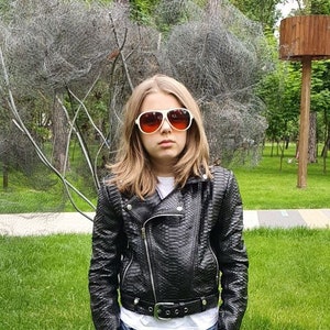 May include: A young person wearing a black leather jacket with a zip fastening, blue jeans, and white sunglasses. The person is standing in a grassy area with trees in the background.
