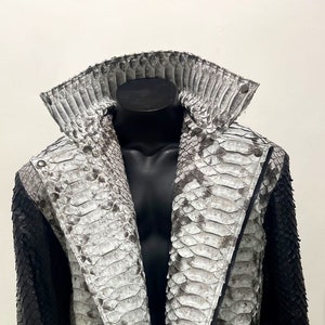 以下が含まれることがあります： ジッパー留めとベルトが付いた黒と白のヘビ革ジャケット。ジャケットはスタンドカラーと長袖です。