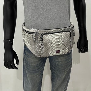 May include: A grey and white snakeskin fanny pack with a zip closure and multiple pockets. The bag is worn around the waist and has a silver-tone zip. The fanny pack is on a mannequin wearing a grey t-shirt and blue jeans.
