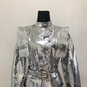 May include: A silver metallic jacket with a high collar, long sleeves, and a belt. The jacket has a snakeskin pattern and a button closure.